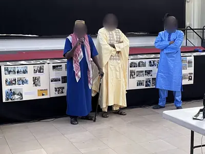 Trois hommes en tenues africaines colorées, debout devant des panneaux d'exposition, celui de gauche tenant un micro.