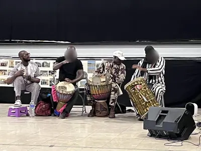 Quatre musiciens assis jouant du djembé et d'autres percussions devant des panneaux de photos.