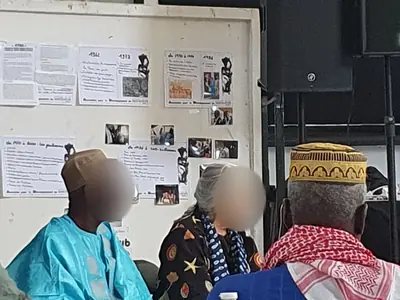 Un homme en boubou turquoise et une femme, de profil devant un panneau d'exposition.