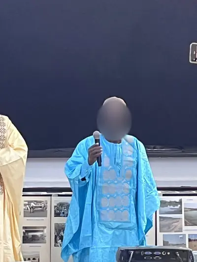 Un homme en boubou turquoise tient un micro et prend la parole, des photos affichées derrière lui.
